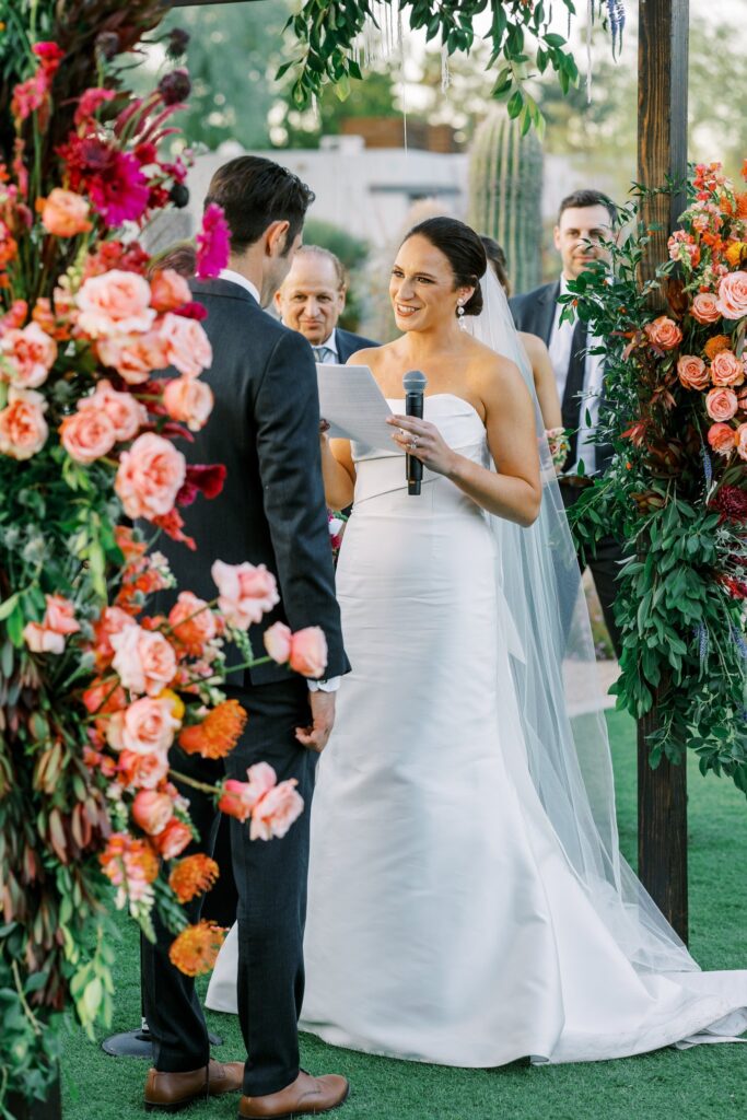 Bride holding microphone and pice of paper, talking to groom under wedding ceremony chuppah.