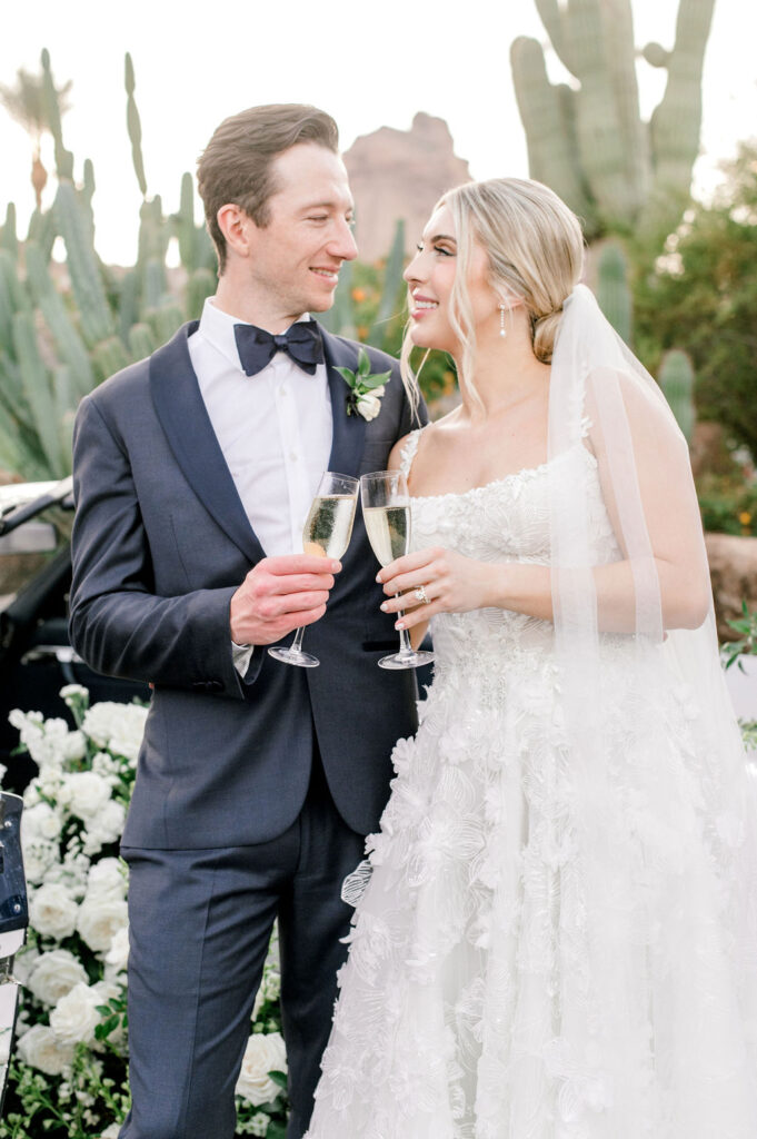 Bride and groom toasting champagne glasses and looking at each other smiling in desert landscape.