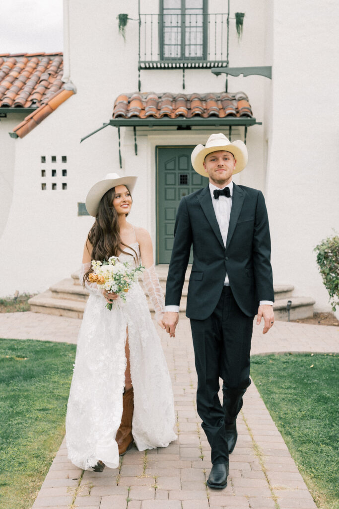 Bride and groom walking down brick path away from house holding hands, bride holding bouquet and looking at groom, both wearing cowboy hats and boots.