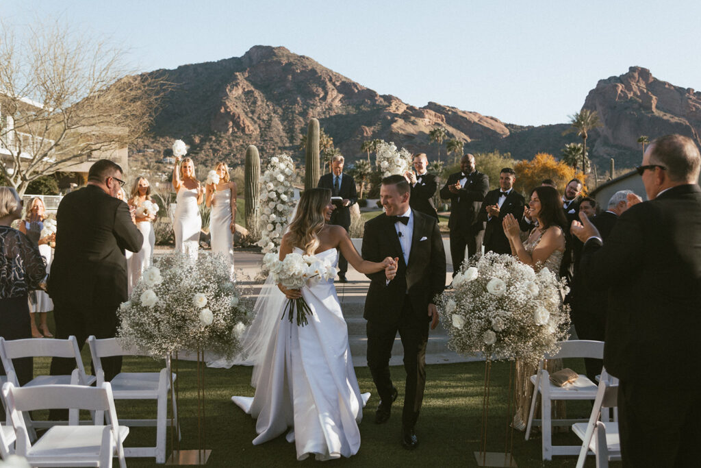 Bride and groom holding hands, exiting wedding ceremony aisle with guests standing and clapping in chairs around them.