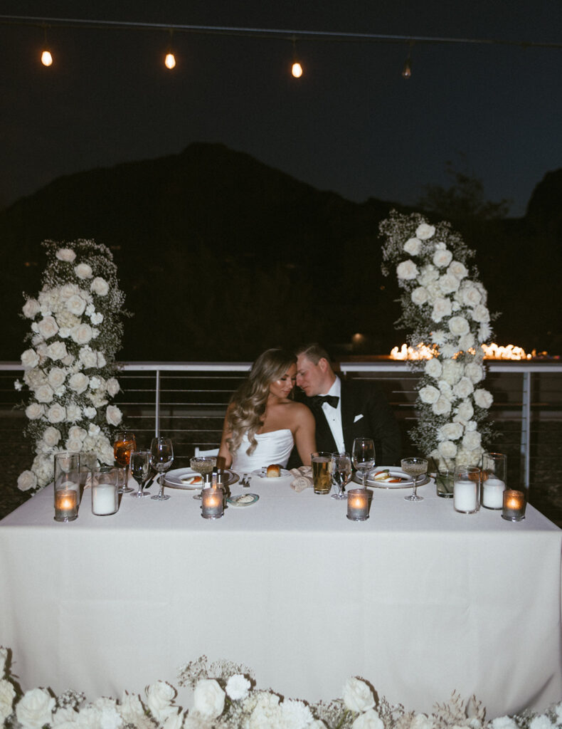 Bride and groom resting heads together while seated at sweetheart table during outdoor reception.