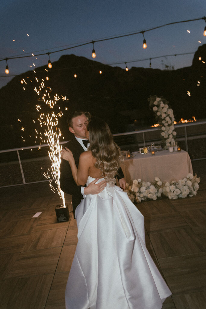 Bride and groom kissing during wedding reception dance with sweetheart table and shooting sparklers behind them.