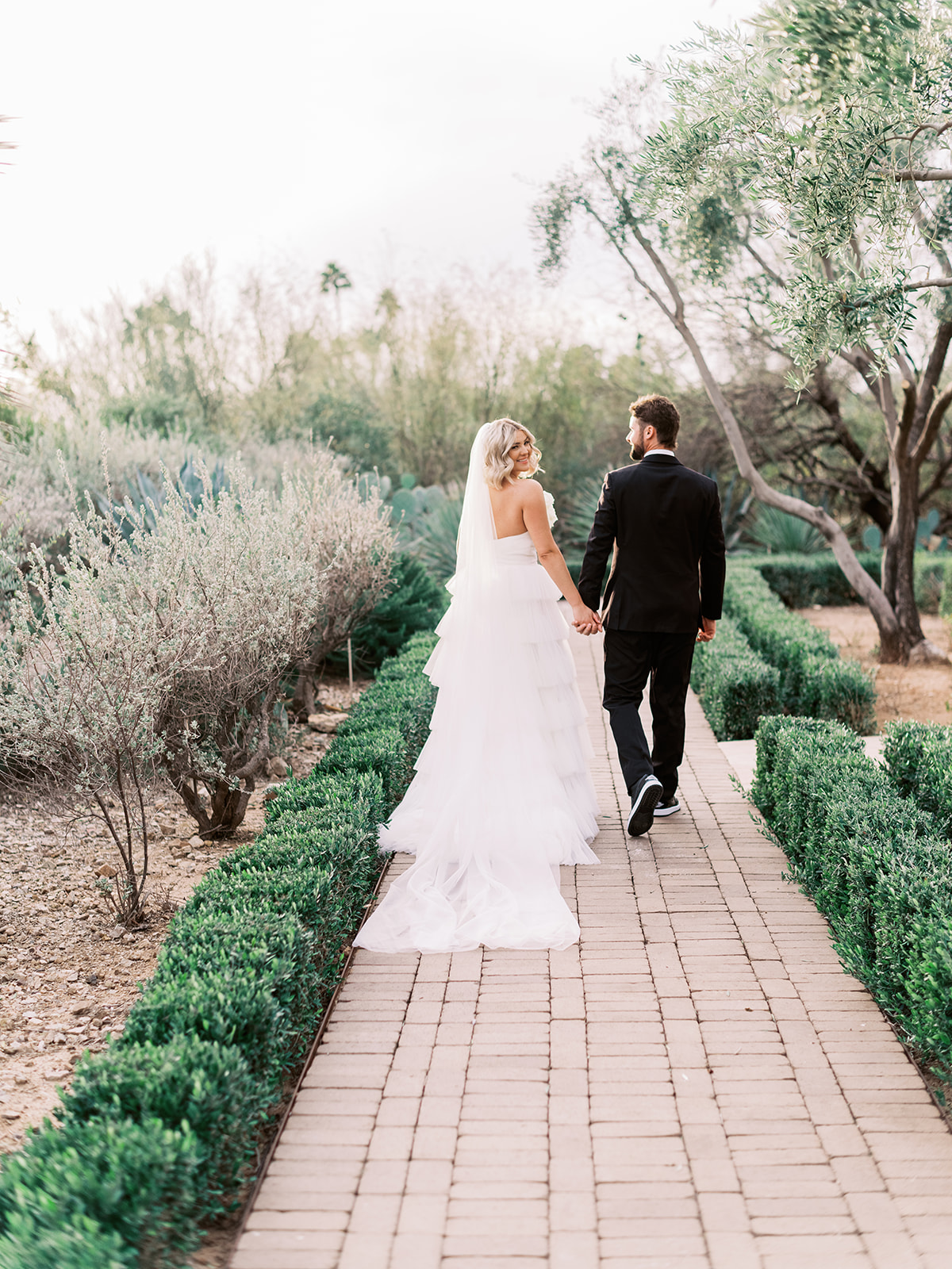Bride and groom walking down brick path holding hands, bride looking back over her shoulder smiling.