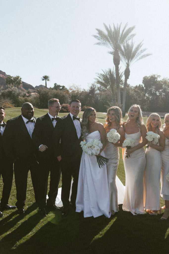 Bride and groom in center of wedding party standing in a line, groomsmen in tuxes and bridesmaids in cream dresses holding white roses bouquets.