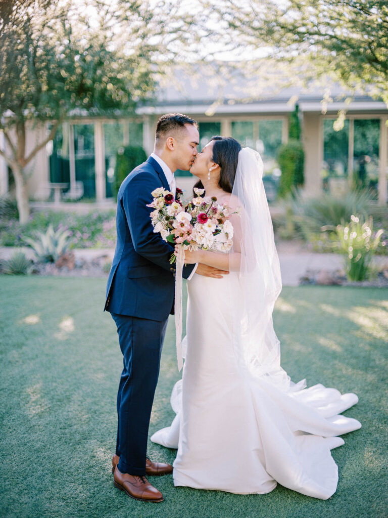 Bride and groom kissing while standing on grass, bride holding bouquet.