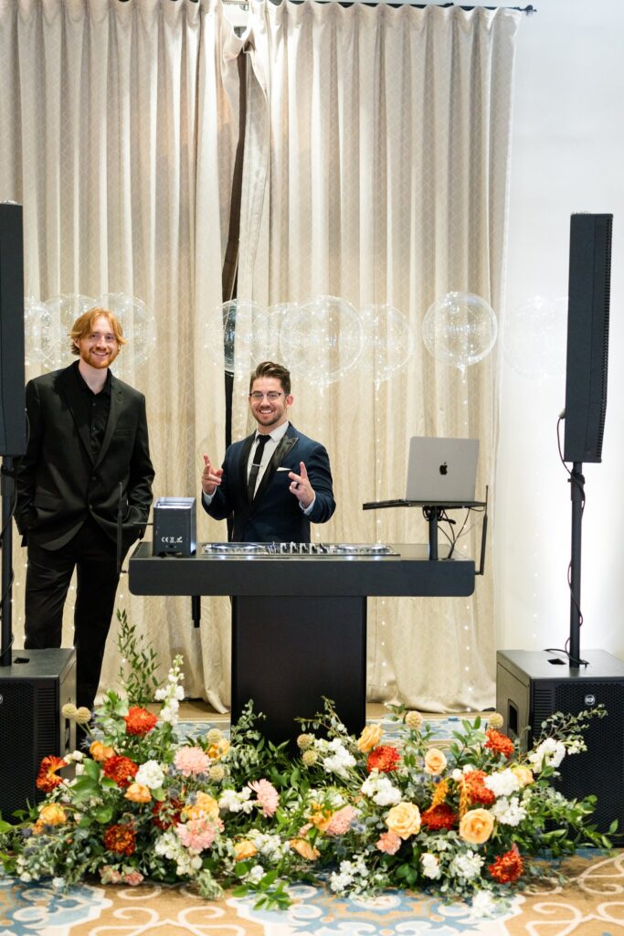 Wedding reception DJs with floral decor placed on ground in front of table.