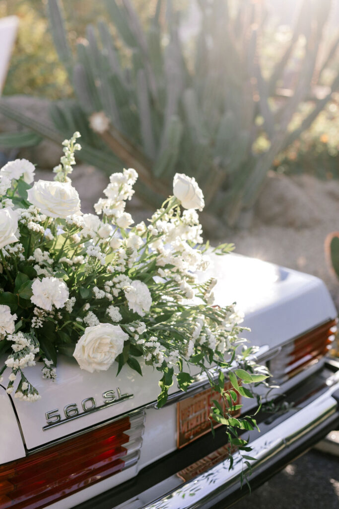 White flowers and greenery arrangement placed on the back trunk of white classic car.