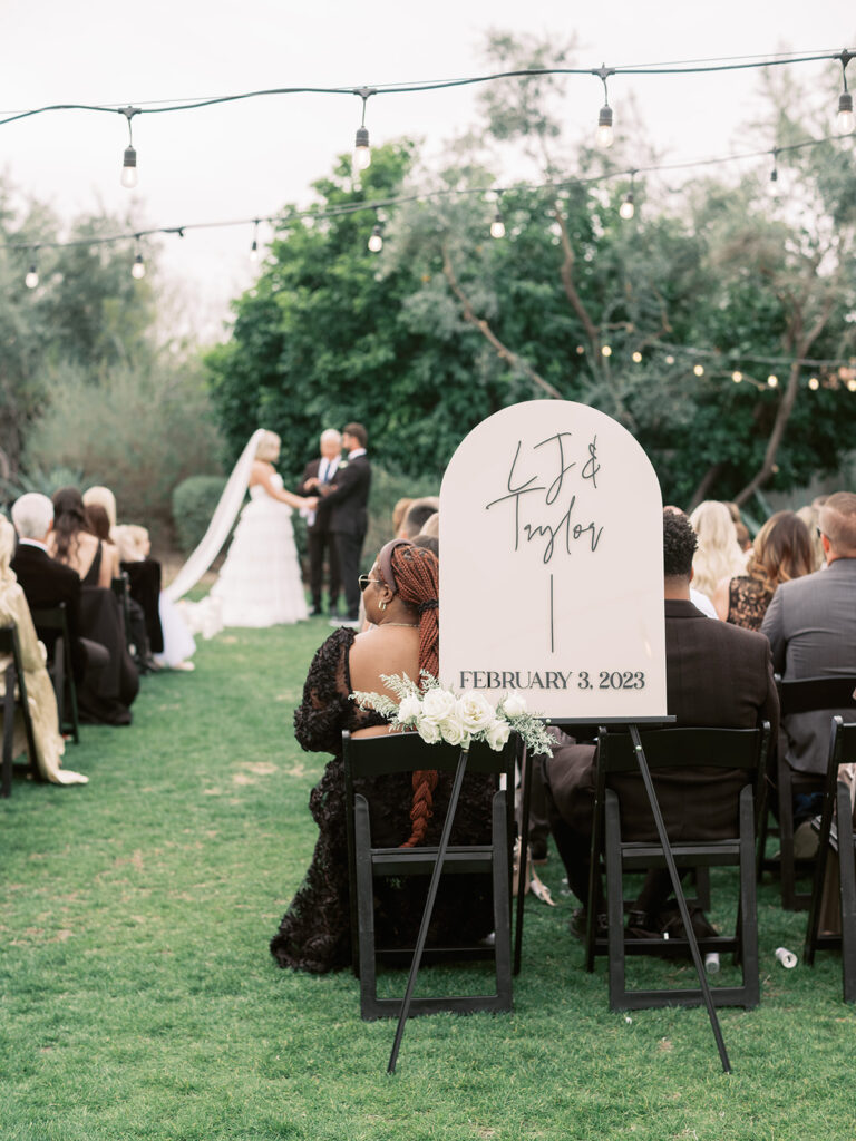 Outdoor wedding ceremony with custom wedding sign behind last aisle with white roses added to it.