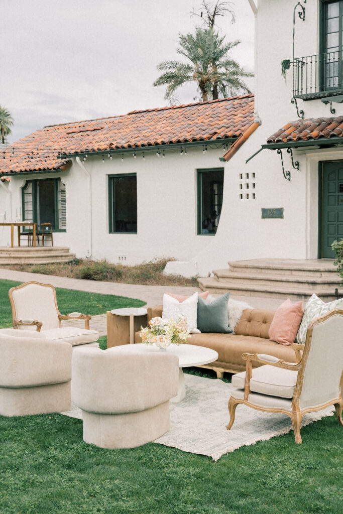 Staged outdoor lounge space with brown leather couch, two arm chairs and two modern rounded chairs. around a coffee table with a floral centerpiece all placed on a rug.