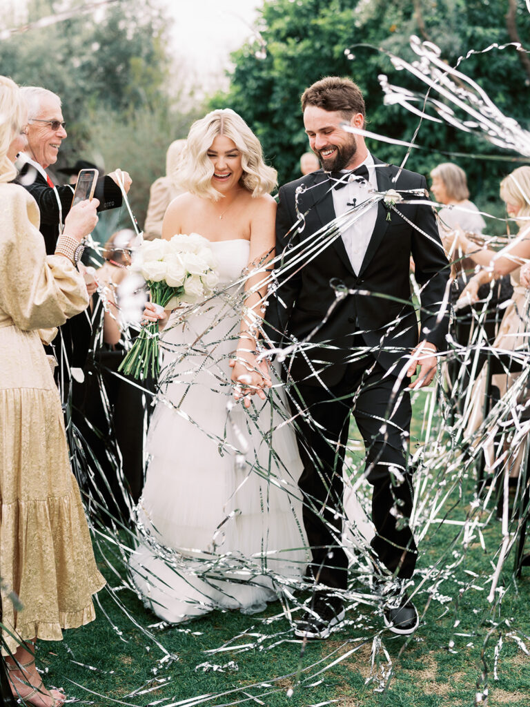Bride and groom walking down wedding ceremony aisle with silver streamers floating around them.