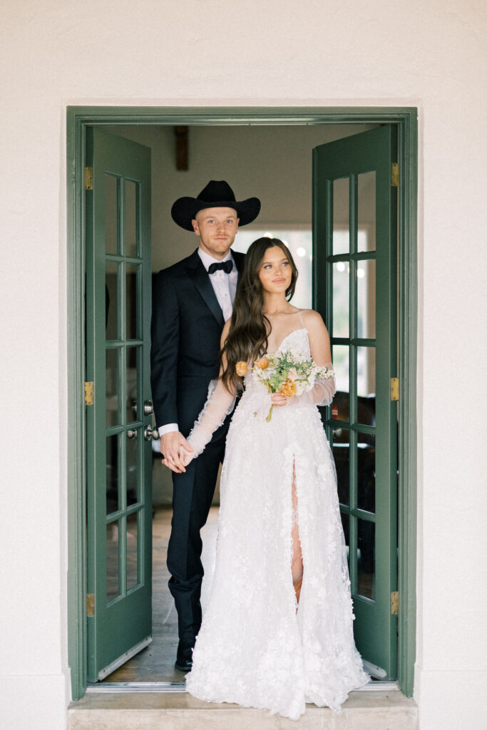 Groom with cowboy hat on standing behind bride holding bouquet in doorway of double green doors opening inside.