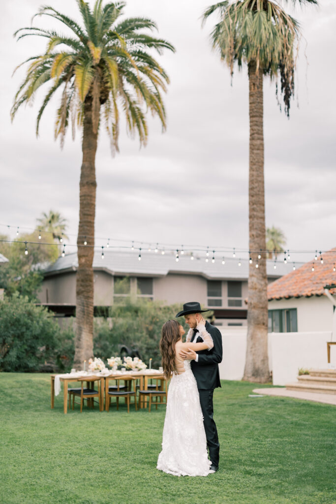 Bride with her hands gently around groom's neck, looking up at him and groom with hands on her back, both standing in grass yard with two tall palm trees behind them.