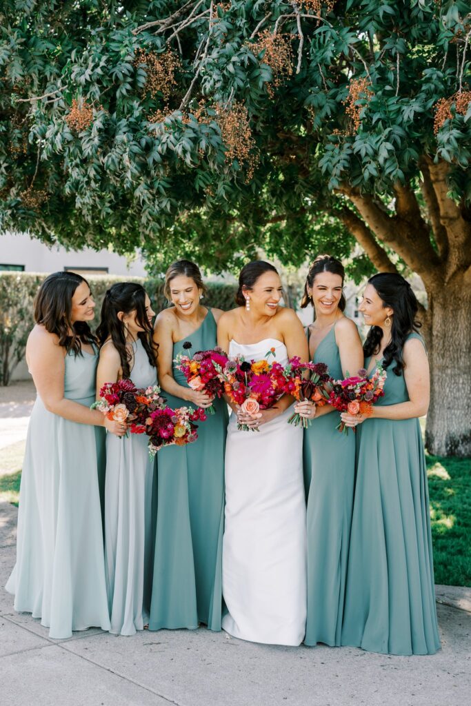 Bridesmaids standing in a line under a tree with bride all holding bouquets and smiling.