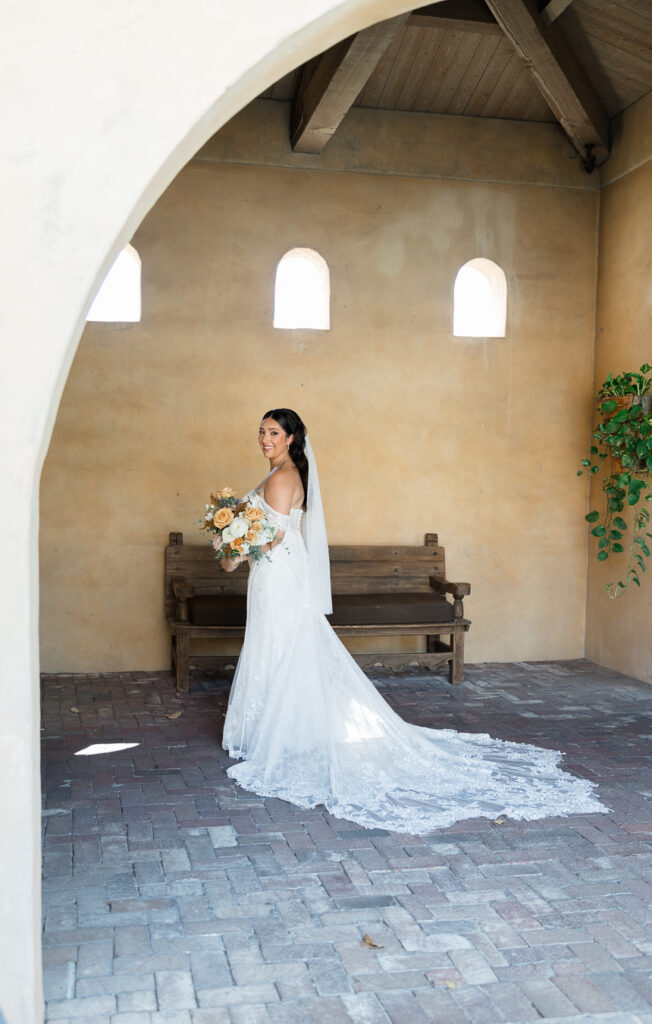 Bride standing in open room, looking over shoulder smiling and holding bouquet.