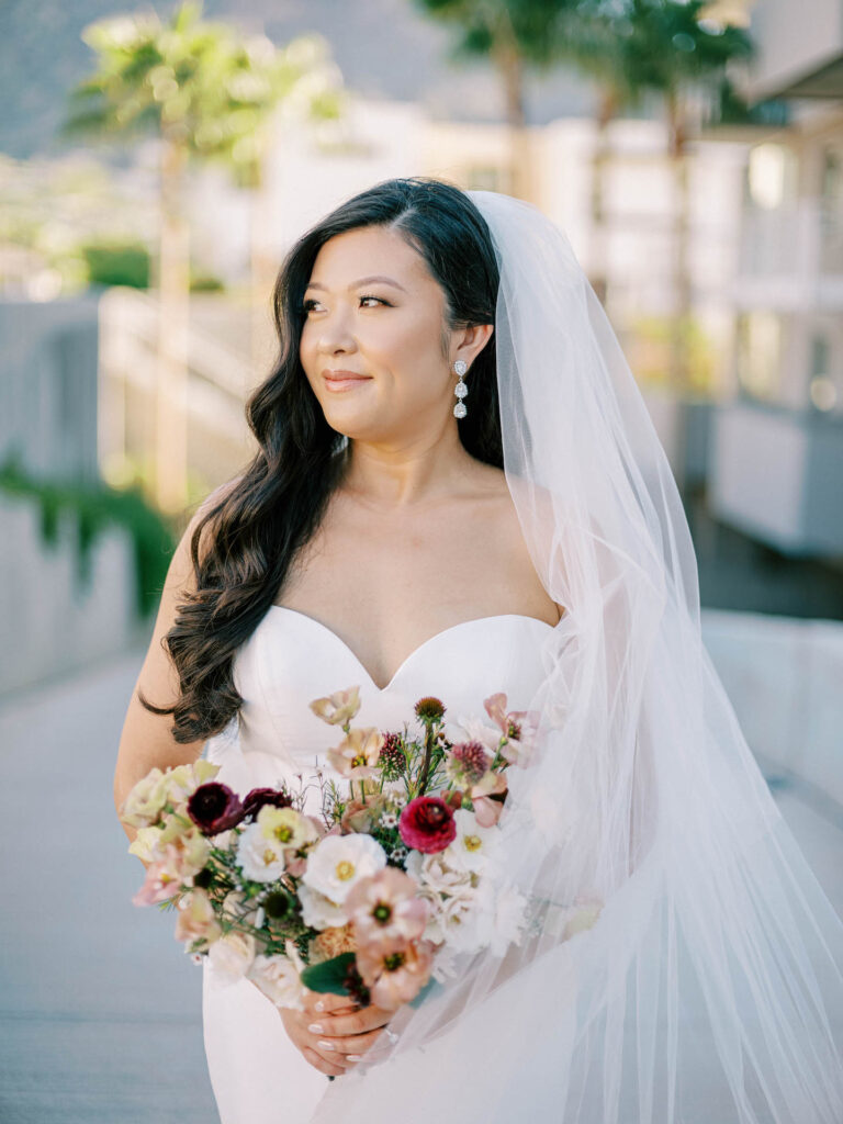 Bride holding bouquet, looking off to side with veil trailing down her other side.