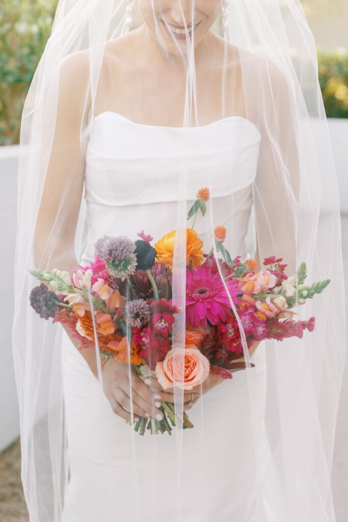 Bride holding bouquet of colorful flower of pink, orange, maroon, and fuchsia behind veil.