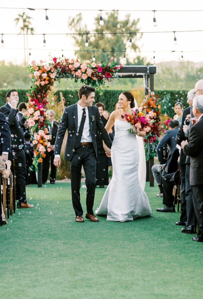 Bride and groom walking down ceremony aisle holding hands and smiling at each other.