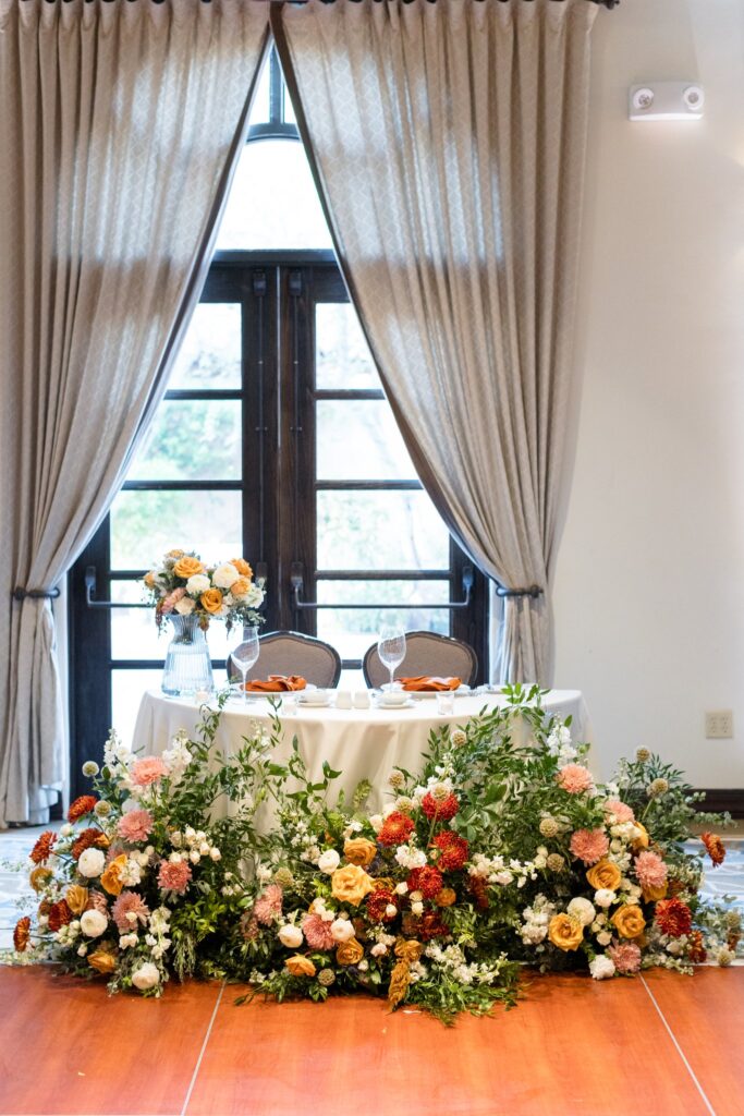 Wedding reception sweetheart table with floral placed in front on ground and arrangement placed on top of table.