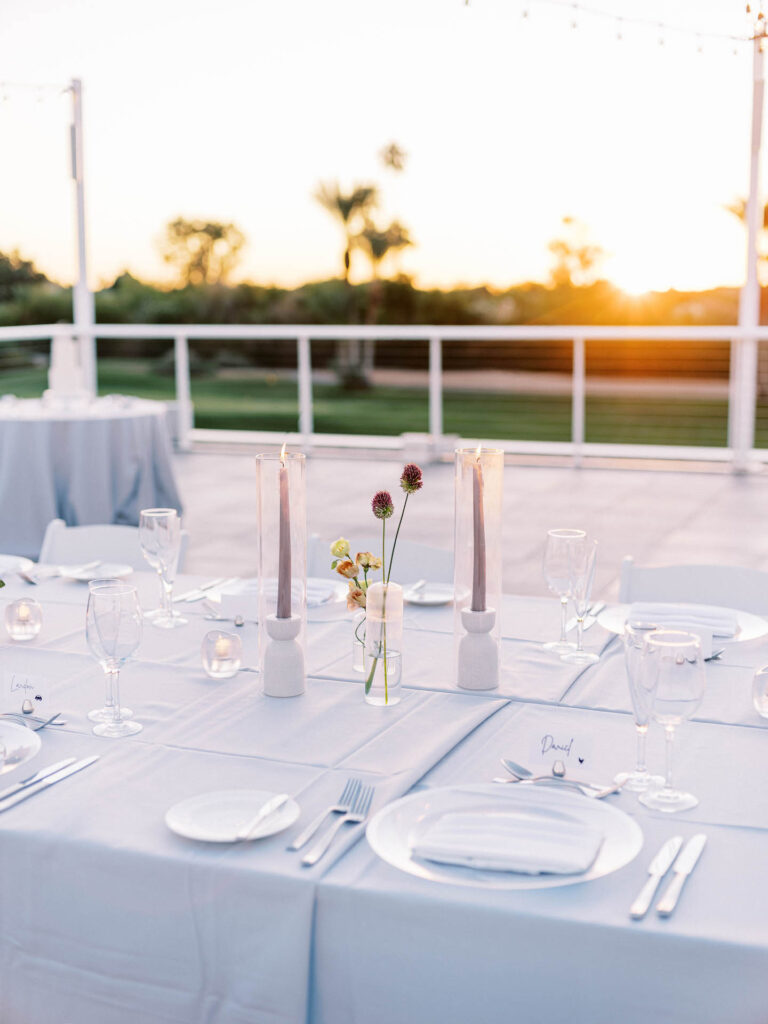 Long reception table place settings with light gray linen, taper candles, and bud vases of floral.