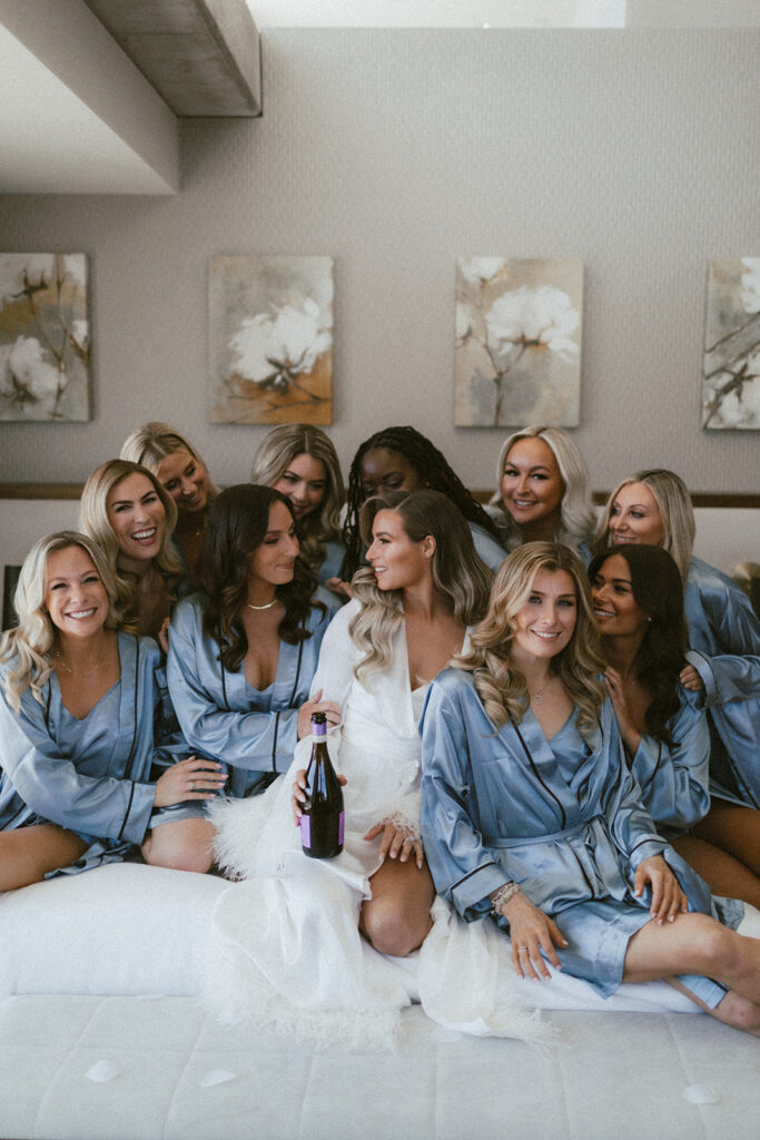 Bride in white robe with bridesmaids in blue robes all sitting on a bed smiling, bride holding bottle of champagne.