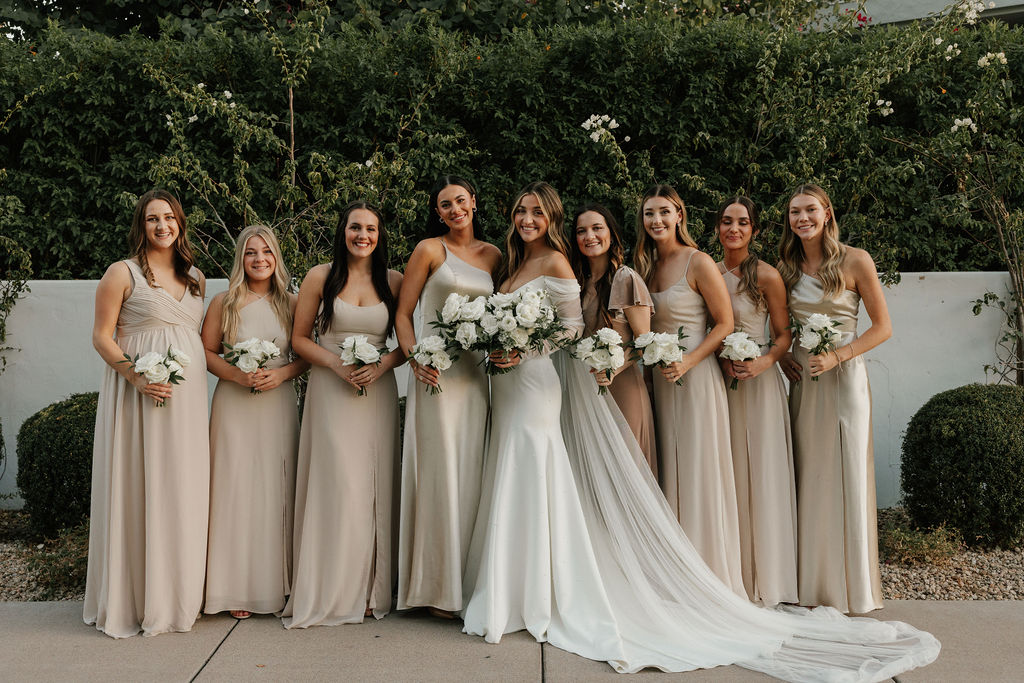 Bride with bridesmaids in taupe dresses, all holding bouquets of white flowers.