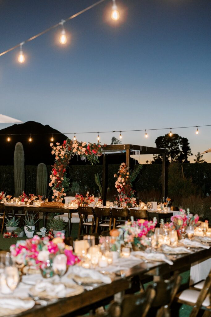 Outdoor wedding reception with long rectangular tables and a chuppah behind tables with floral installed on it.