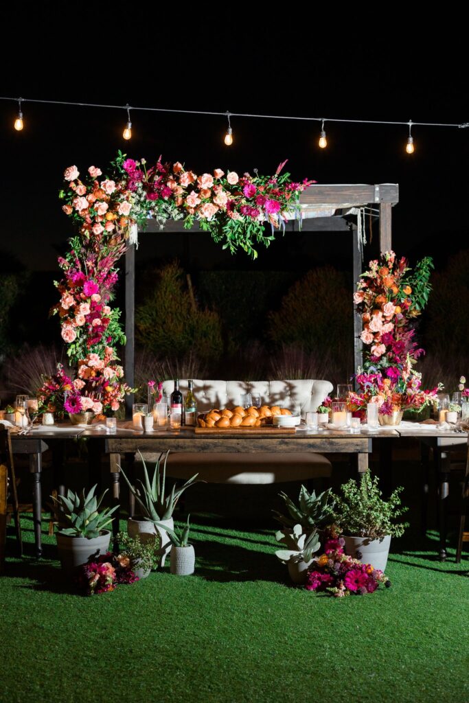 Sweetheart table with plants and floral on ground in front of it, flower arrangements on the surface, and installed floral behind on chuppah.