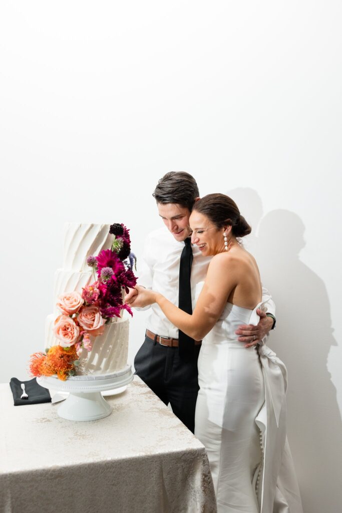 Bride and groom cutting three tiered white wedding cake with floral added to front.