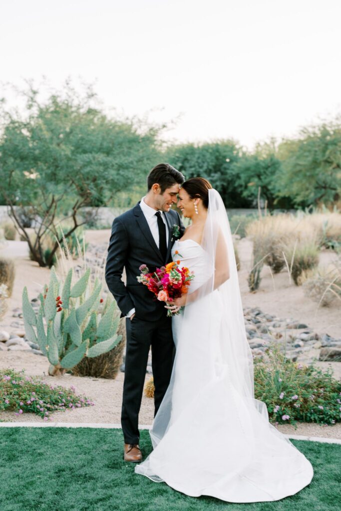 Bride and groom standing on grass in front of desert landscape, smiling and resting foreheads together.