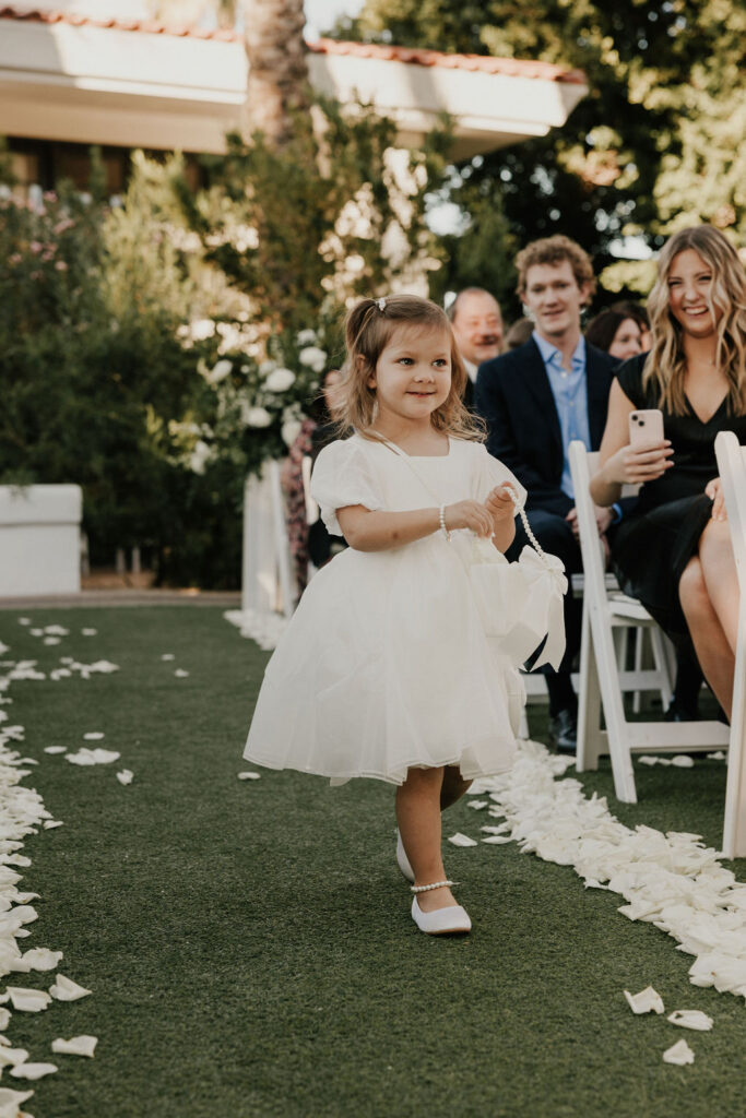 Flower girl in white dress walking down aisle, smiling.