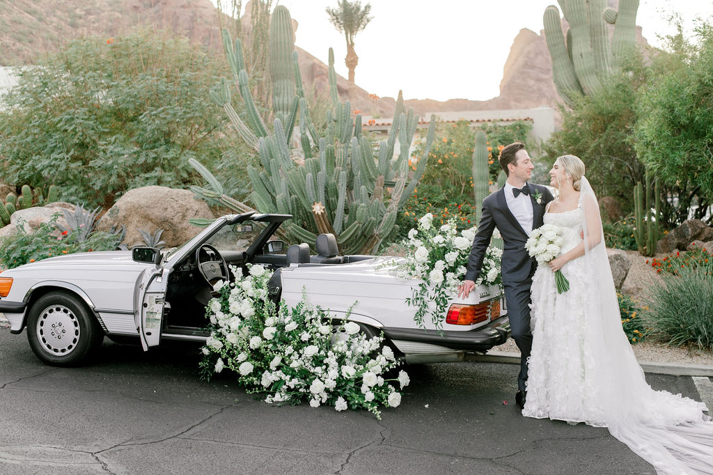 Bride and groom standing behind vintage convertible car smiling at each other with white flowers and greenery floral arrangements placed on trunk and flowing out of driver's side open door.