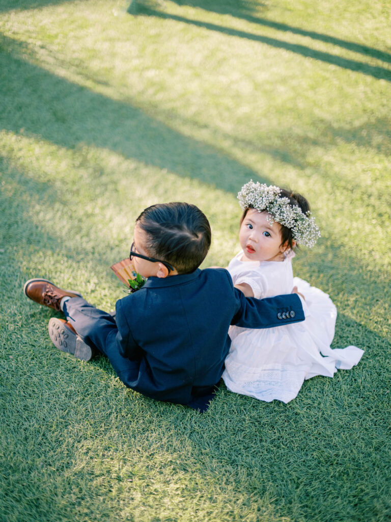 Ring bearer and flower girl seated on grass, with ring bearer having arm around flower girl and flower girl looking up.