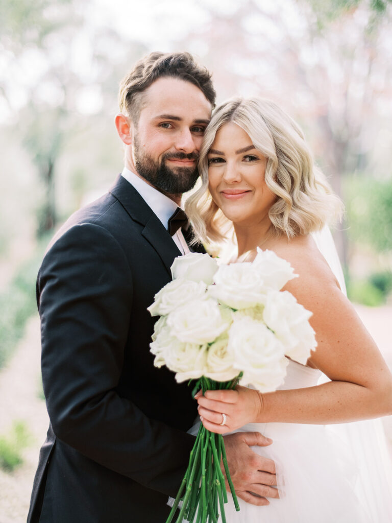 Bride and groom embracing, smiling, bride holding bouquet of long stem roses.