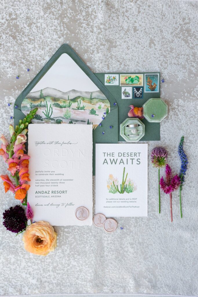 Desert themed wedding invitation flat lay in green and white with live floral details around it and wedding rings.