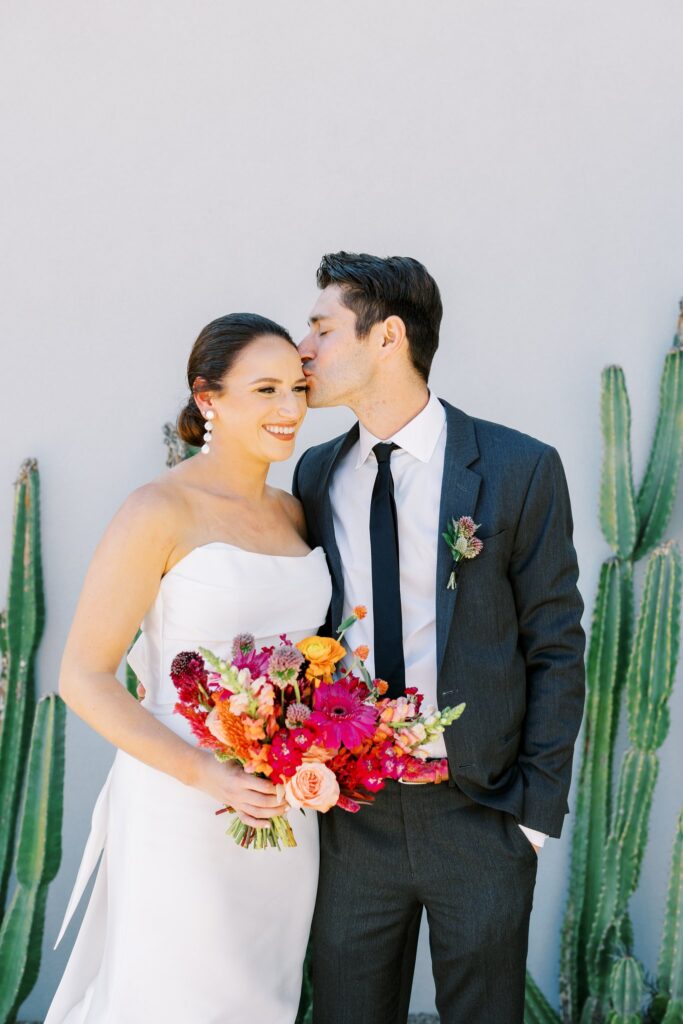 Groom kissing top side of bride's head and bride smiling off to side, holding bouquet.