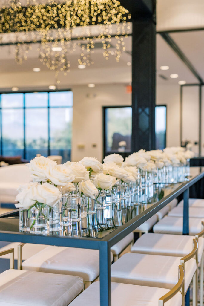 Indoor glass table with many glass bud vases with white roses in them on top.