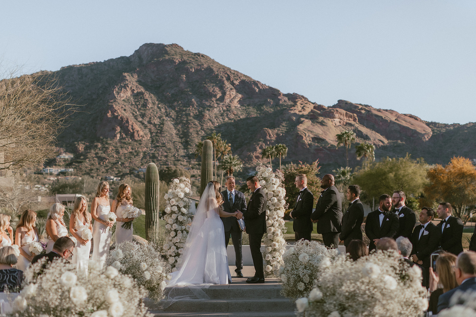 Bride and groom standing in ceremony altar space at Mountain Shadows with Camelback Mountain in background, bridesmaids and groomsmen on either side and officiant behind them.