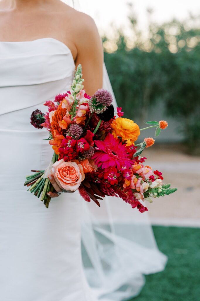 Bride holding out bouquet of pink, orange, maroon, and fuchsia flowers.