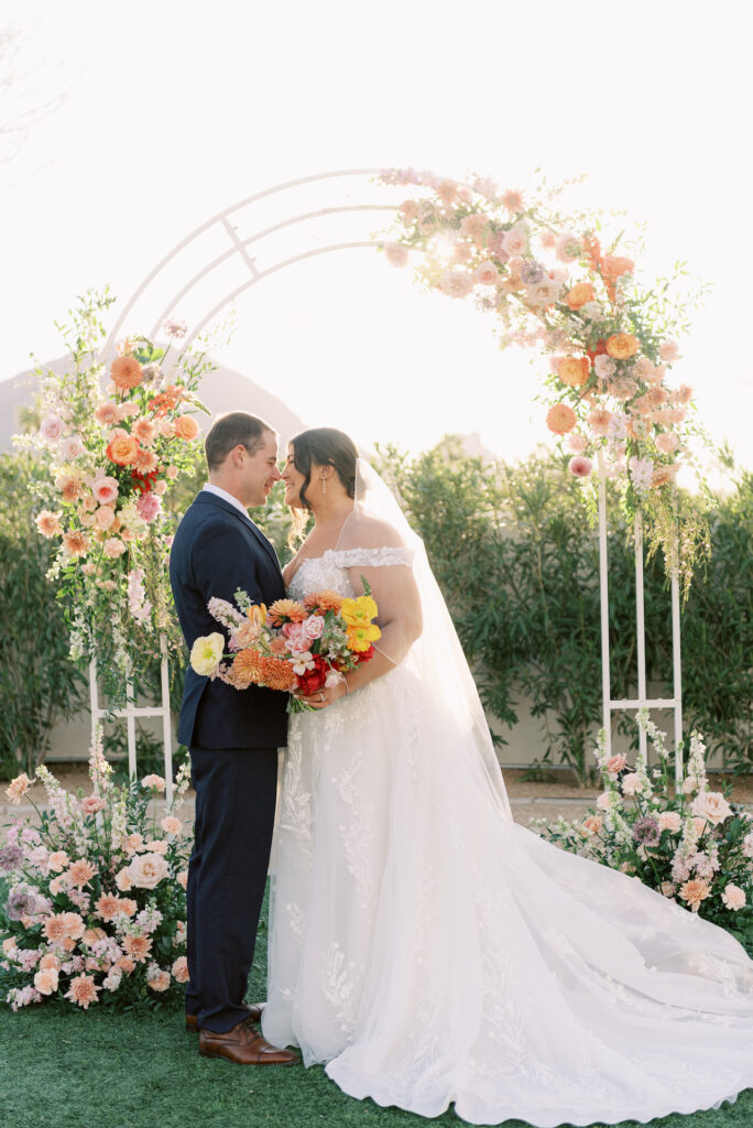 Bride and groom in front of wedding ceremony arch, smiling with noses touching and flowers in pink, peach, and orange on arch, ground and in her bridal bouquet.