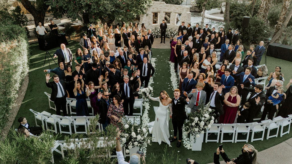 Bride and groom at the end of the wedding ceremony aisle smiling up with all the guests for picture taken from up above.