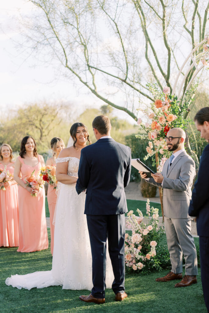 Bride and groom during wedding ceremony standing in altar space, holding hands, back of groom and can see bride smiling with wedding party on either side and officiant.