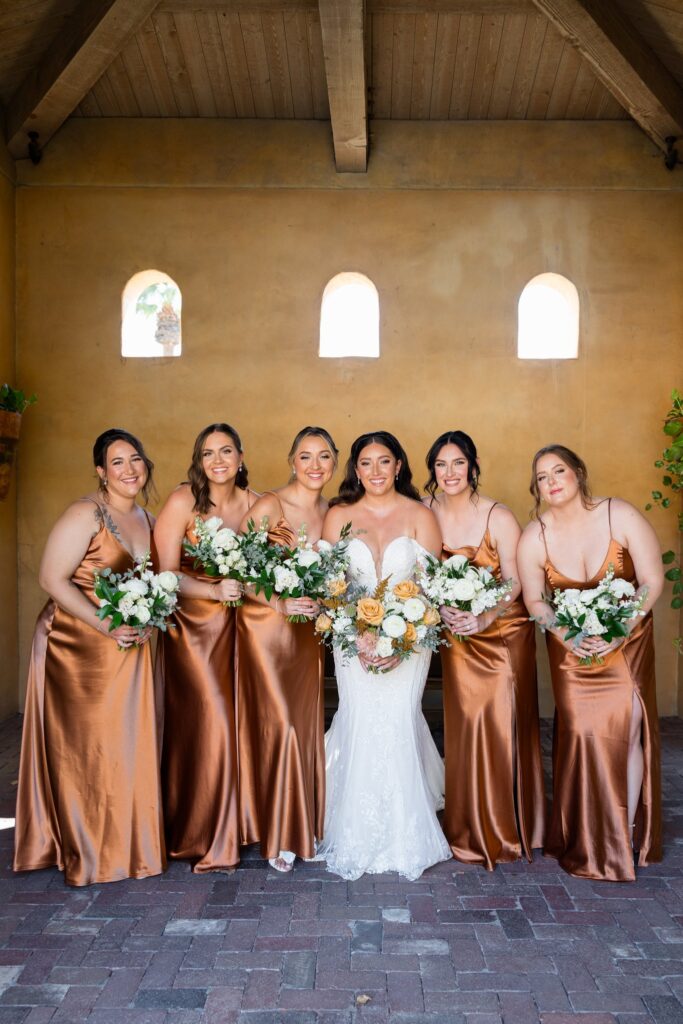 Bride standing in a line with bridesmaids in copper colored dressed all holding bouquets under building roof.