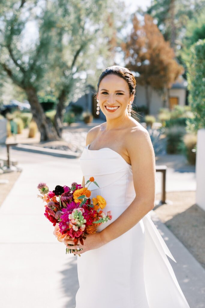 Bride smiling, holding bouquet of bright flowers, standing on paved path.