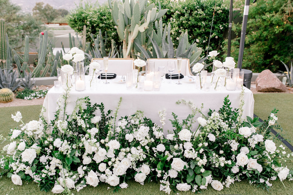 Sweetheart table with white roses bud vases and candles on top and white flowers and greenery ground arrangements in front.