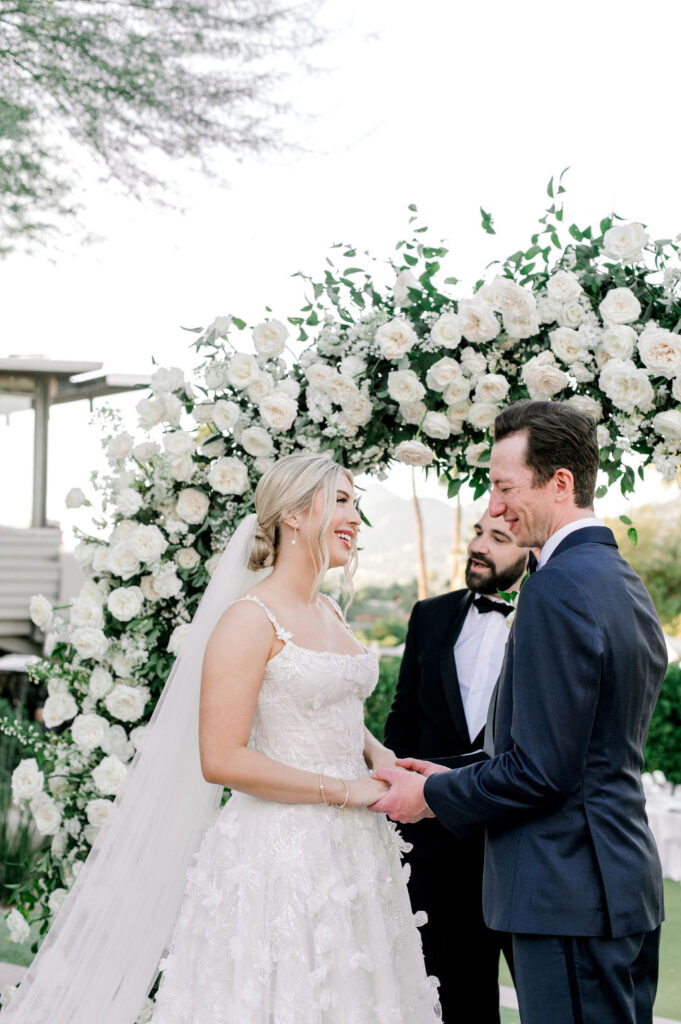 Bride and groom holding hands under white roses floral arch during wedding reception.