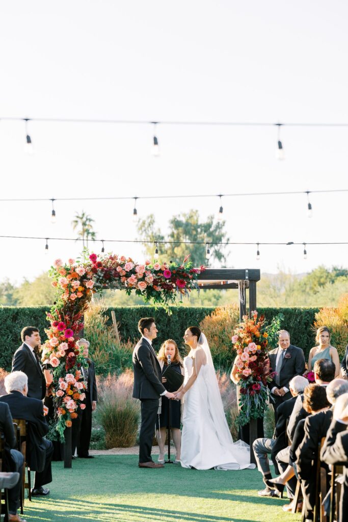 Bride and groom holding hands under wedding chuppah in ceremony altar space with officiant behind them.