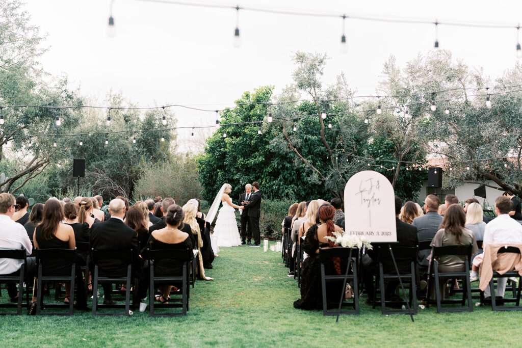 Outdoor wedding ceremony with guests seated in black folding chairs and couple in altar space with officiant.