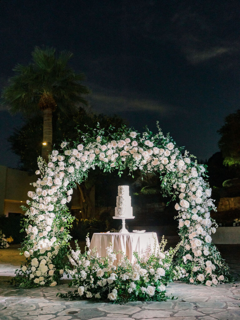 Wedding cake table with three tiered white cake on stand and an arch of white flowers and greenery behind it.