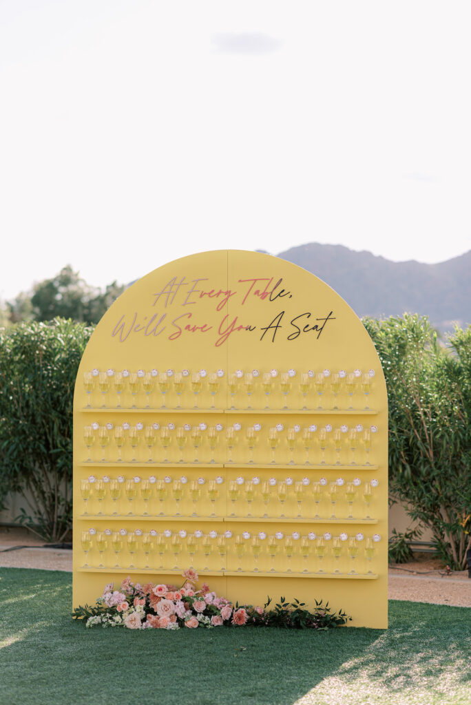 Custom yellow wedding reception escort board with shelves of champagne glasses.