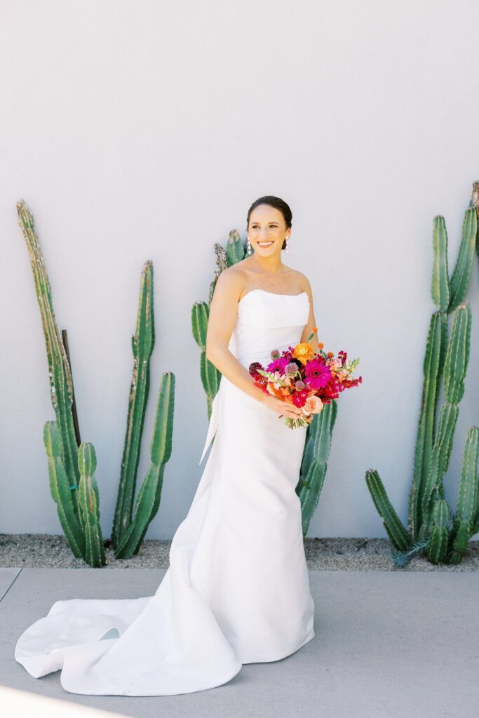 Bride smiling off to side, holding bouquet down at waist, standing in front of white wall with cactus growing in front of it.